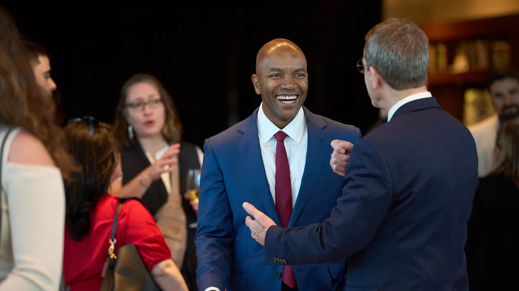 Dr. Michael Johnson in navy blue suit and maroon tie smiles warmly while conversing with another man at an impact summit, with guests visible in background.